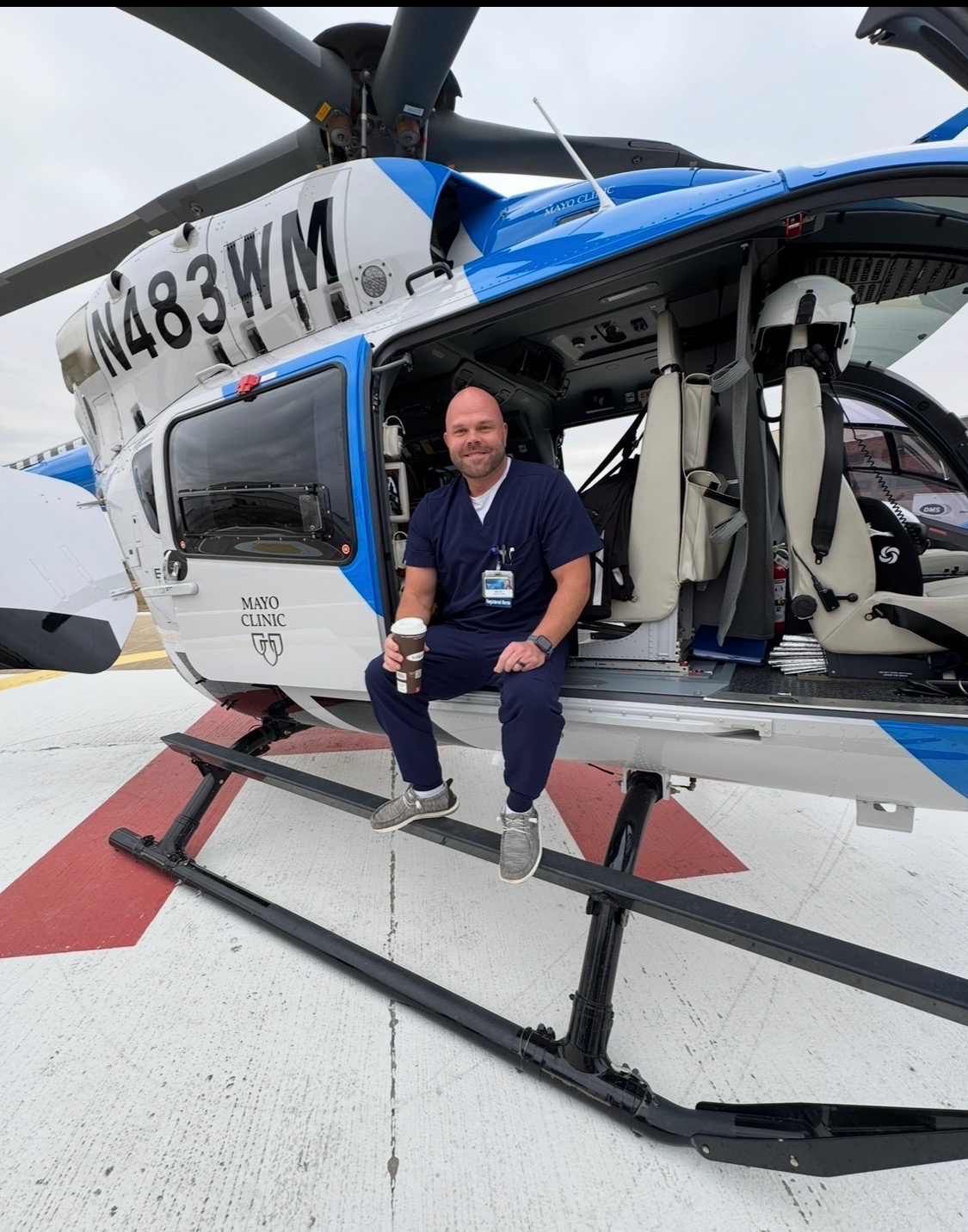 Nick in dark blue scrubs sitting on the open door of the Mayo 1 medical transport helicopter (N483WM), holding a coffee.