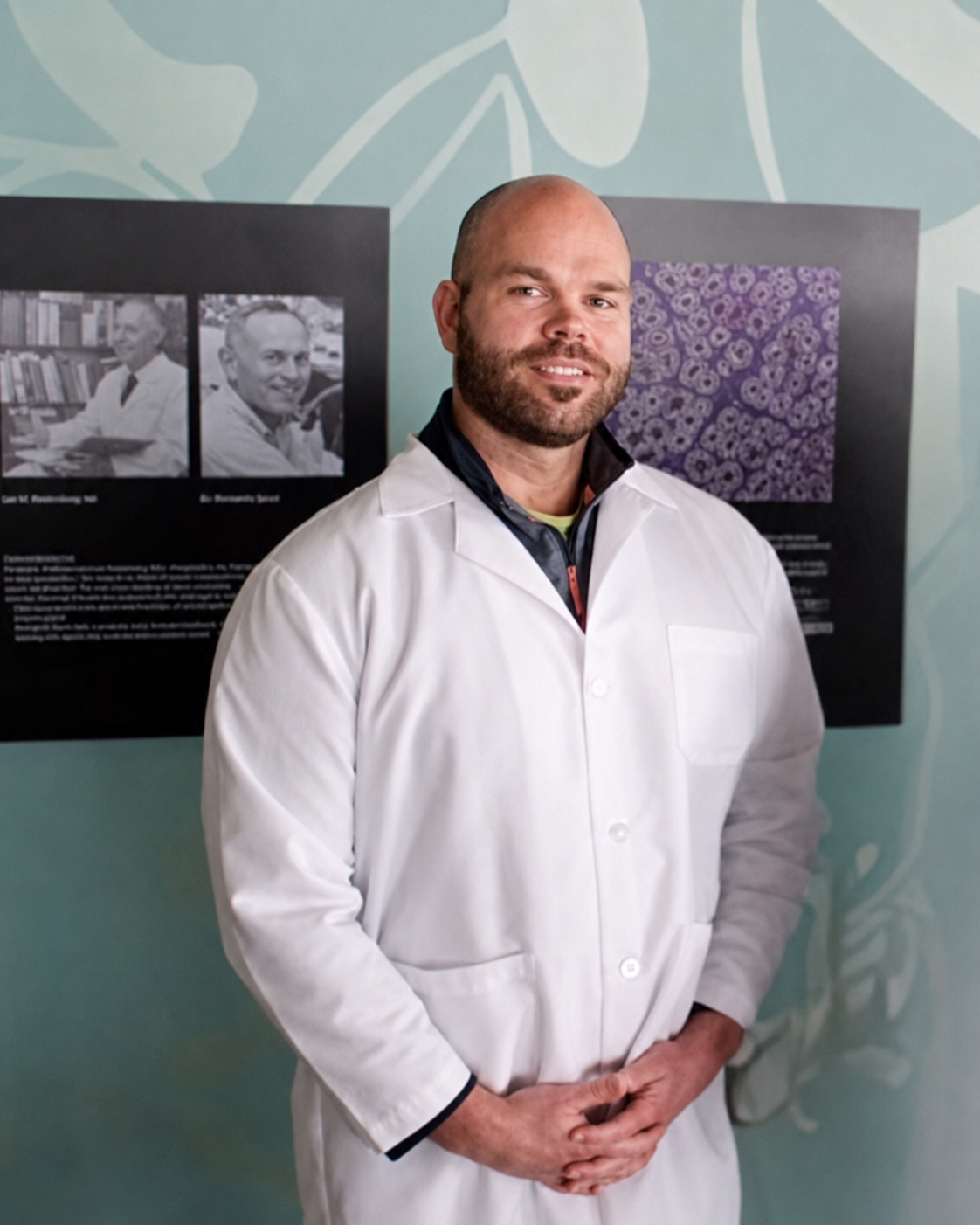 Nick in a white lab coat at the University of Minnesota Hormel Institute, where he ran translational oncology research on cancer epigenetics. The framed portraits behind him are of Ralph Holman and colleagues who discovered omega-3 essential fatty acids at the same institute.